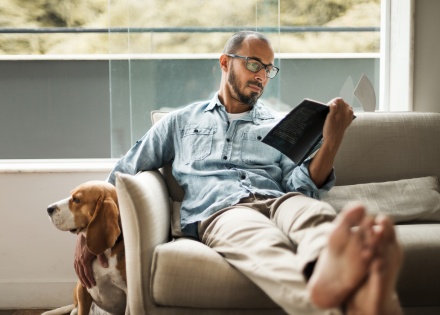 Man with his feet up on a couch, reading a book and petting his dog