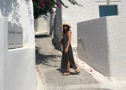 Jenny Ackerman, a woman with fair skin and long brown hair, standing in sunlight among white Grecian-style buildings.