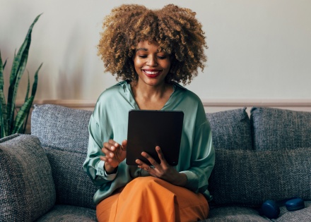 A woman sitting on a couch at home, smiling while browsing for Rippling alternatives for payroll and EOR solutions