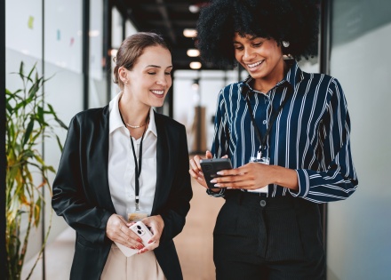 Two smiling colleagues looking at a phone in an office, discussing OTE salary.