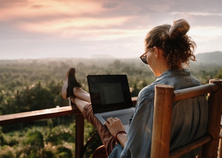 Woman working remotely from a balcony overlooking a lush landscape of trees