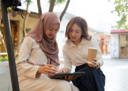Two women sitting outside among a courtyard looking at and speaking about something shown on a tablet