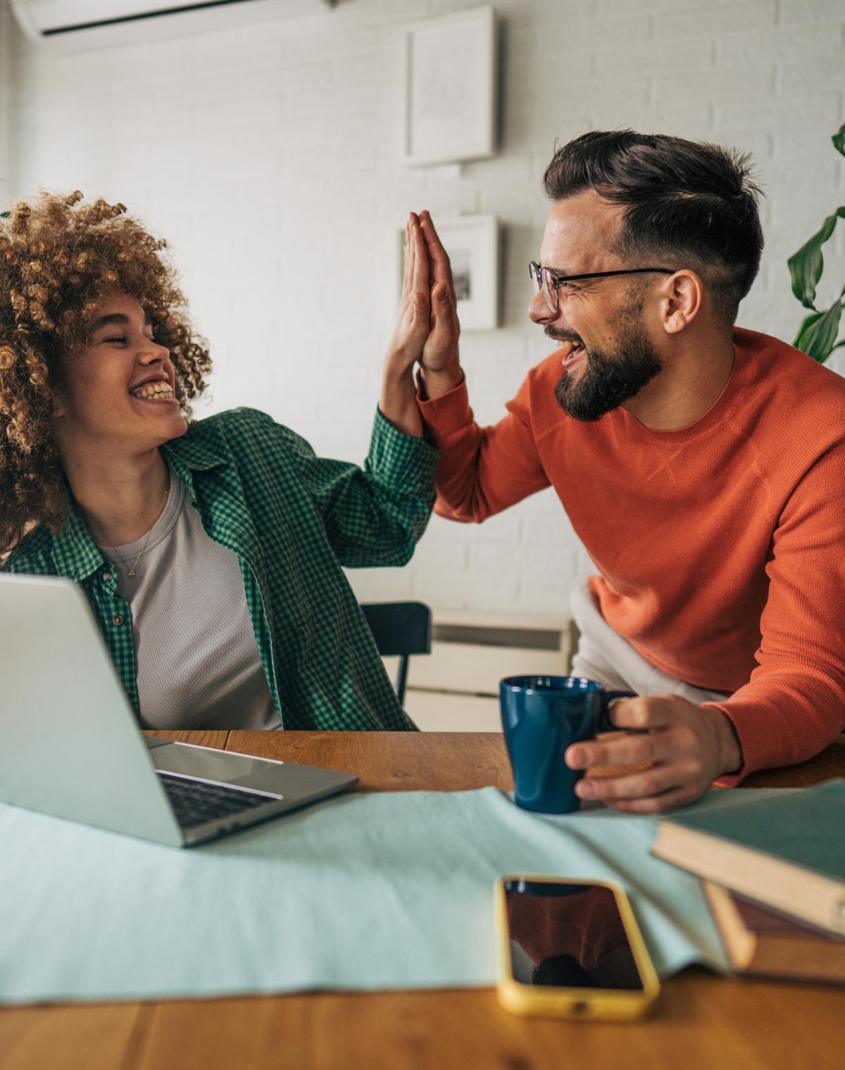 Man holding coffee high-fives his female colleague as they sit at a table in front of laptop.