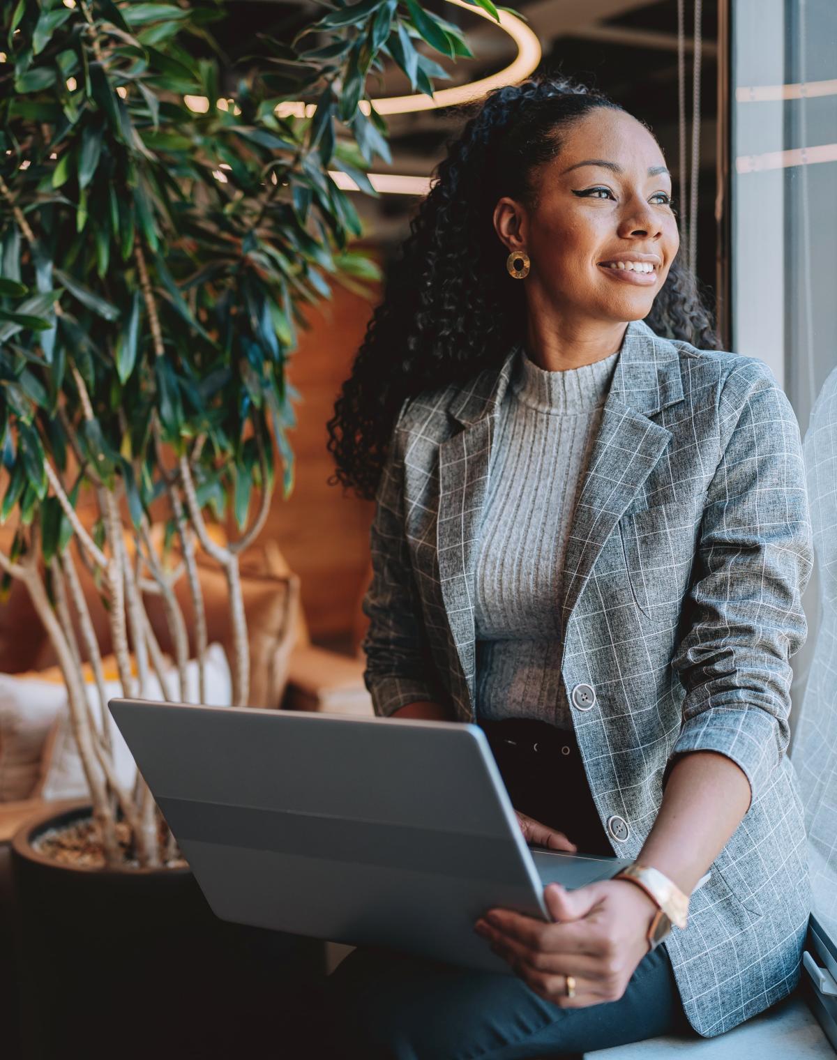 A professional woman smiles while looking out the window while holding a laptop.