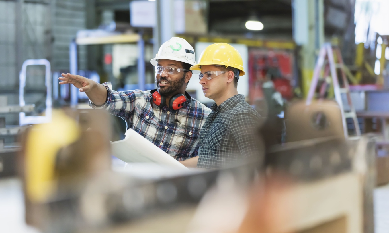 Two colleagues in hard hats and safety glasses reviewing building plans.