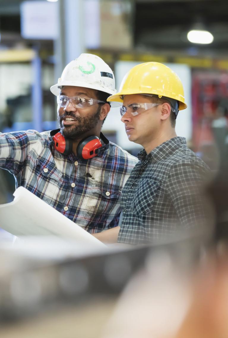 Two colleagues in hard hats and safety glasses reviewing building plans.