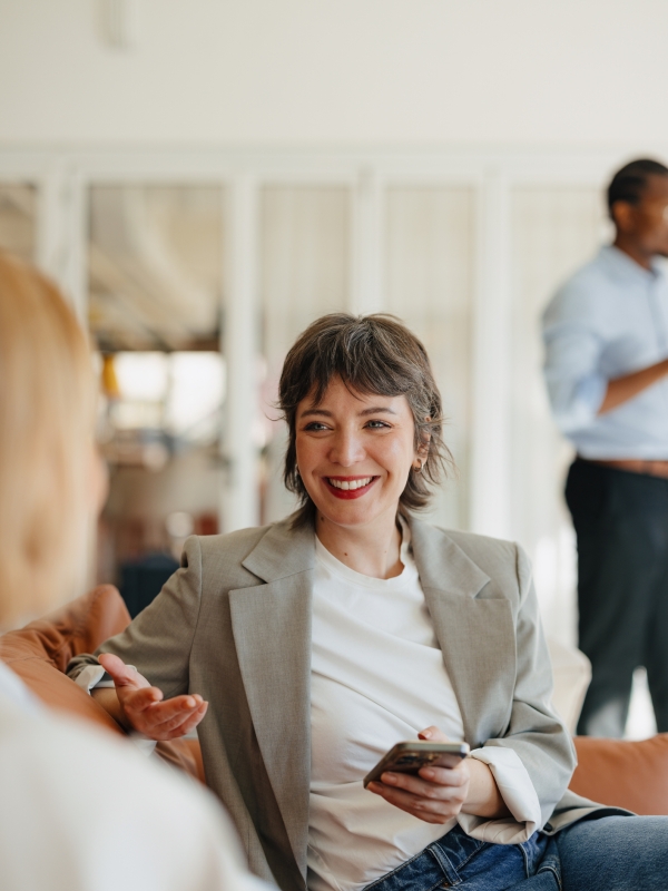 Woman in business casual attire holding phone and discussing M&A with colleagues.