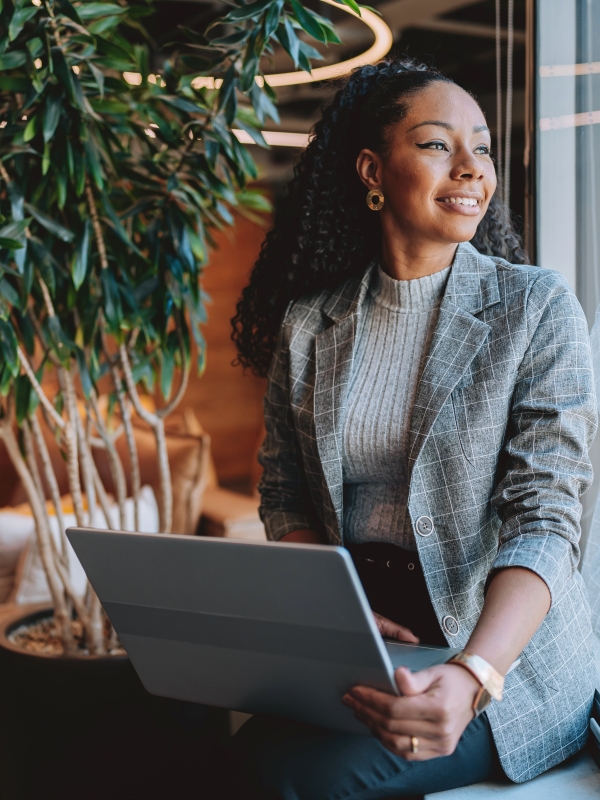 A professional woman smiles while looking out the window while holding a laptop.