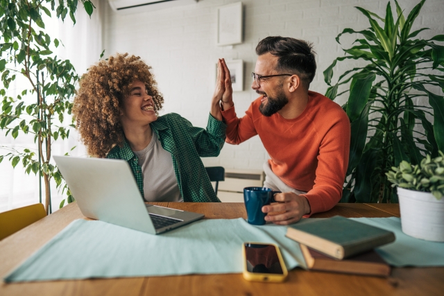 Man holding coffee high-fives his female colleague as they sit at a table in front of laptop.
