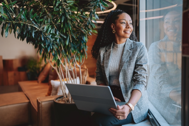 A professional woman smiles while looking out the window while holding a laptop.