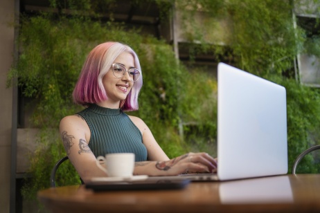 Female employer with pink hair sitting at outdoor table working on laptop with coffee.