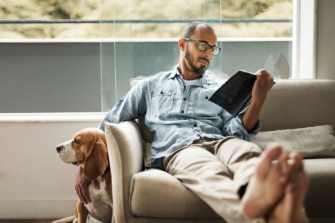 Man with his feet up on a couch, reading a book and petting his dog